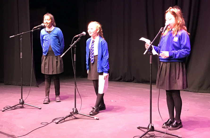 Photo of 3 children standing on a stage in front of microphones